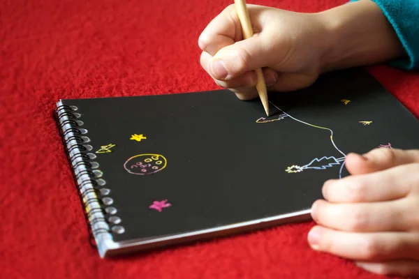 Child\'s hand draws a landscape drawing in a notebook with black paper. Close up process of drawing by the child in album with black pages. Children\'s creativity