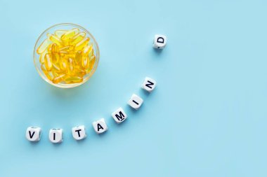 Yellow capsules in the round glass bowl and the word vitamin D from white cubes with letters on a blue background. VITAMIN D word for healthy and medical concept. Sunshine vitamin health benefits clipart