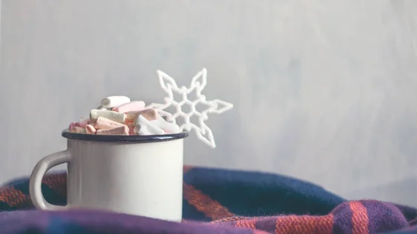 Winter cozy concept. Coffee with marshmallows and decorative shiny snowflake in white enameled metal cup in purple warm wool scarf. Warm weekend in cold weather.