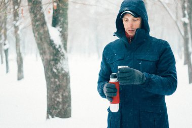 Kış parkında kar lı termos tutan mutlu adam. Genç adam kışın ormanda sıcak içecek içiyor