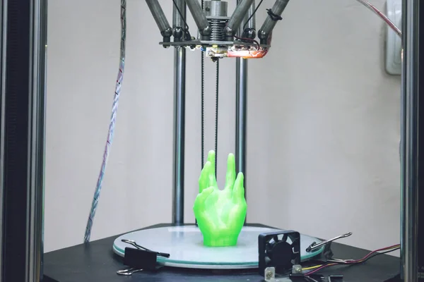 Green plastic 3D printed human hand stands in three-dimensional printer. Object model printed on automatic three dimensional 3d printer. Additive manufacturing and robotic automation technology.