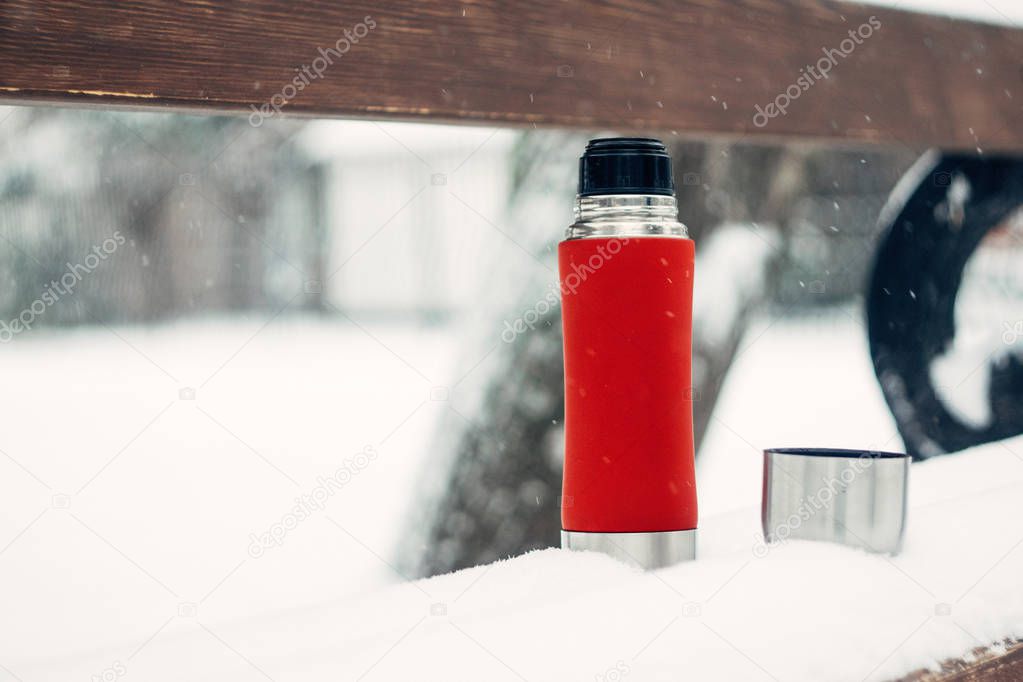 Winter Activities outdoor. Winter holidays, hot drinks concept. Red thermos and cup on snowy bench in winter park. Thermos with hot tea, coffee, mulled wine, glogg