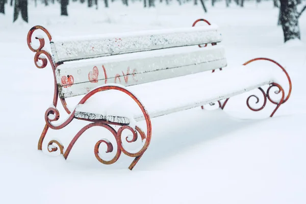 Snow-covered bench with heart symbol in the city park. Forged metal and wooden park bench and trees covered by heavy snow. Winter date, romance, Valentine\'s Day,