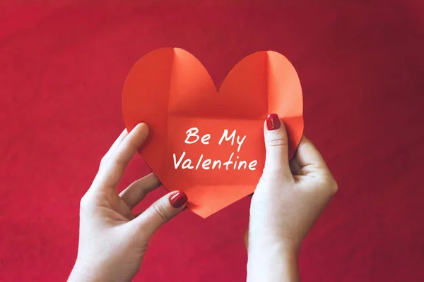 Valentines day card ideas. Greeting card in womens hands. Female hand with heart shape card with text Be my Valentine on red background. Love, romance concept