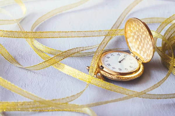 Gold Vintage pocket watch with golden ribbons on grey cement background. Hourglass or sand timer, symbol of time. Selective focus.