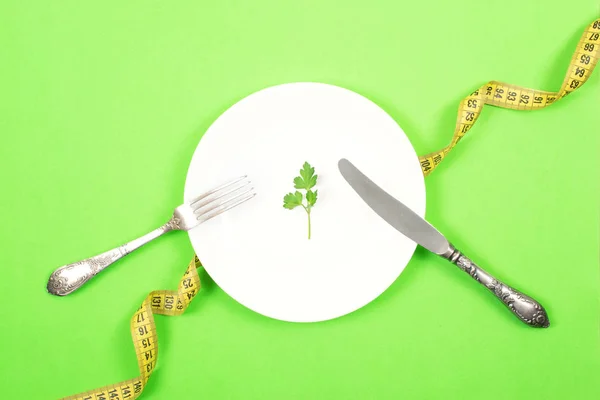 Diet, weigh loss, healthy eating, fitness concept. Small portion of food on big plate. Small green salad leaf on white plate with fork and knife on the background of measuring tape.