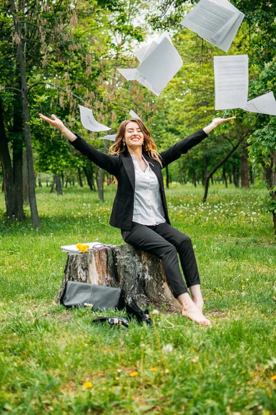 Out of office, dismissal, freelance, distant work, working remotely. Young business woman in suit sitting on grass in park throwing office documents and enjoys freedom