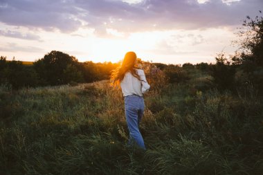Ulusal Rahatlama Günü, gevşeme uygulamaları, akıl sağlığı, yavaş yaşam konsepti. Uzun rüzgarlı saçları ve çiçekli buketi olan genç kız günbatımının arka planında doğanın tadını çıkarıyor..