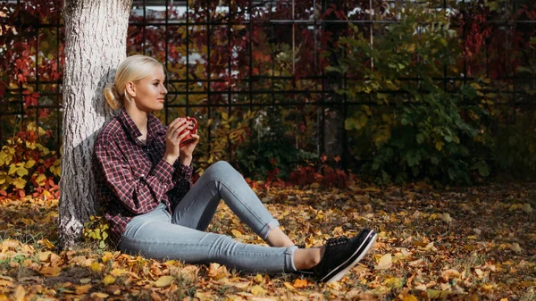 Fall cozy mood concept. Autumn woman drinking coffee on park under fall foliage. Beautiful young modern woman sitting by fall tree and enjoying nature.