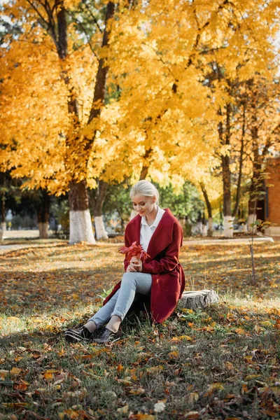 Beautiful blonde Woman with Autumn Leaves on Fall Nature Background. Alone brooding woman in red coat holding autumn leafs in the nature.