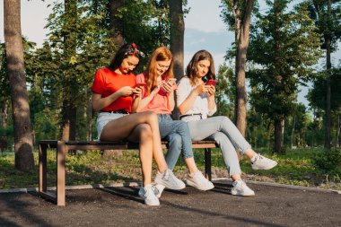 Parkta akıllı telefonlarıyla sohbet eden üç kız. Gen Z genç kız arkadaşları alet kullanıyor ve dışarıda eğleniyorlar..