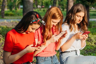 Parkta akıllı telefonlarıyla sohbet eden üç kız. Gen Z genç kız arkadaşları alet kullanıyor ve dışarıda eğleniyorlar..