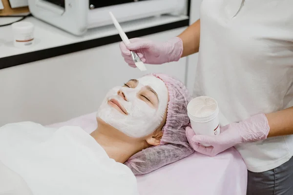 Professional Esthetician Apply Face Mask to the client\'s face in spa beauty center. Young woman getting facial care by beautician at spa salon Acne Treatment, face peeling mask.