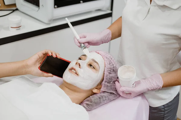 Professional skin care in beauty salon. Young woman with clay face mask takes selfie on phone. Facial care by beautician at spa salon. Acne Treatment, face peeling mask, spa beauty treatment.