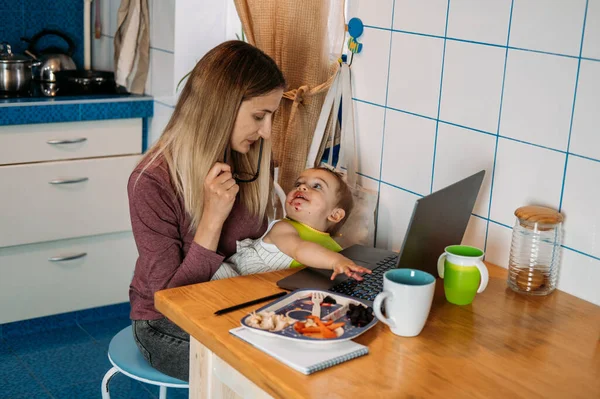 Work from home. Young mother with baby girl working at home using laptop on kitchen background Young woman feeding her baby, talking on mobile phone, looking at laptop at her home working place.