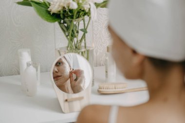 Beyaz havluyla yatak odasında dinlenen ve aynanın yanında kilden maske yapan genç bir kadın. Kız güzellik tedavisi yapıyor ve evde dinleniyor. Sabah cilt bakımı güzellik rutini, kişisel bakım