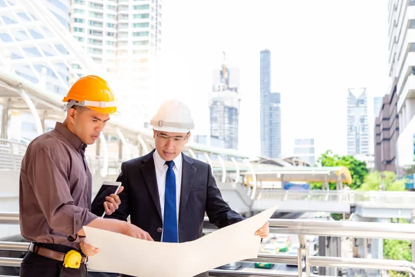 Employee is consulting his engineer boss for solving the problem on blueprint. Handsome engineer man is looking the blueprint. Boss is  thinking and solving the problems with cityscape background