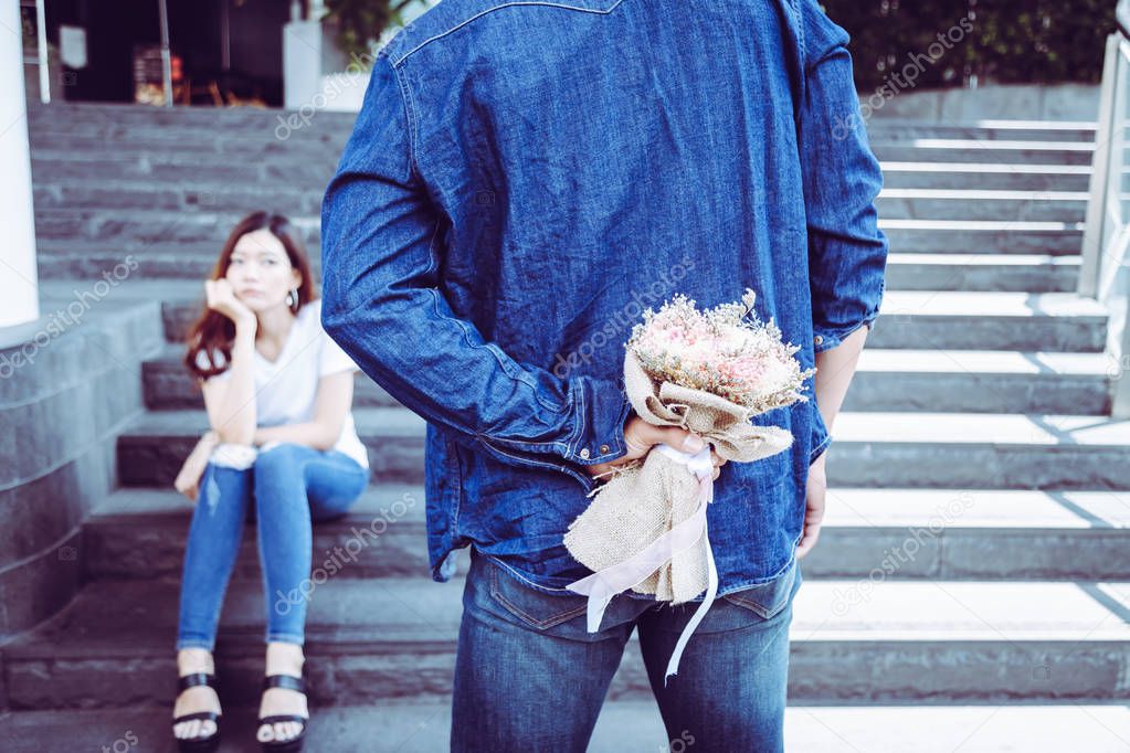 Handsome boyfriend is hiding bouquet of flower behind his back. Attractive handsome guy meet her today to make up with his beautiful girlfriend. His girlfriend is sulking and guy ask for apologize him