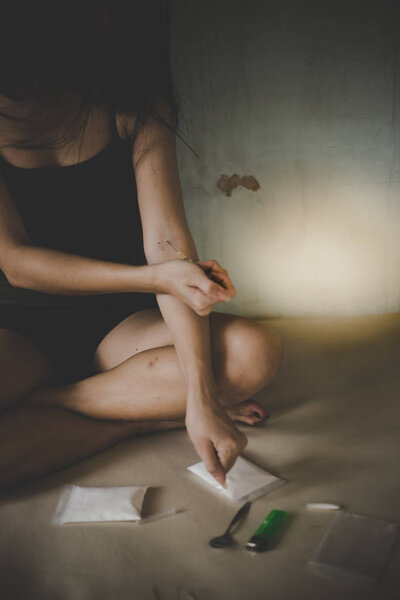 Drug addict woman is injecting heroin, cocaine into arm veins. Poor girl sit on old bed at dark room. Narcotic is unhealthy for body. She is junkie person with sunlight background and copy space