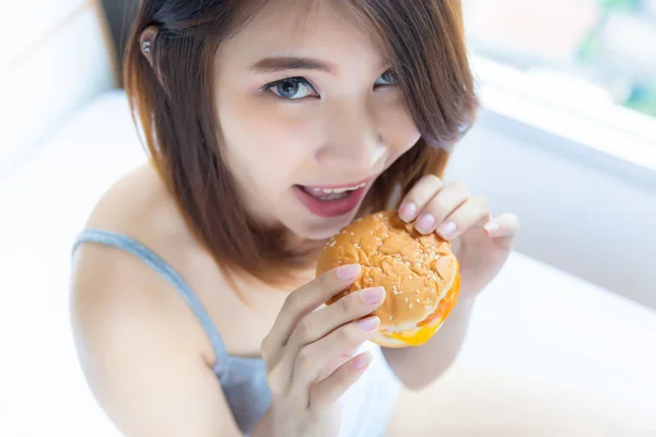 Charming beautiful woman is eating hamburger in the morning on bed. The hamburger has trans fat and unhealthy food. The trans fat is one of cause of illness or cancer. She love eating junk food
