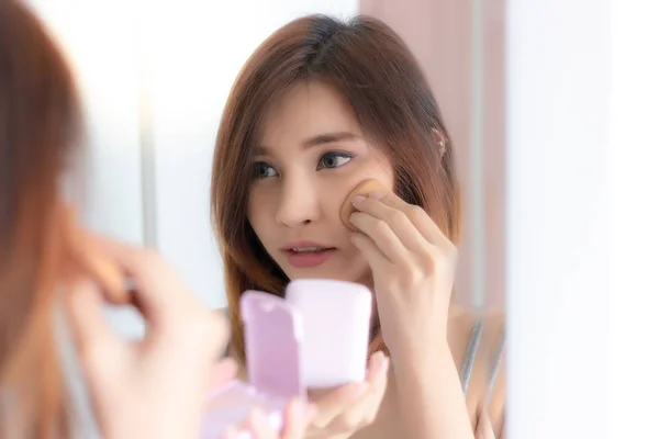 Beautiful Asia woman apply or puts on makeup to her beautiful face for getting nice and shiny skin face by using press powder foundation. Attractive beautiful women looks at the mirror at home