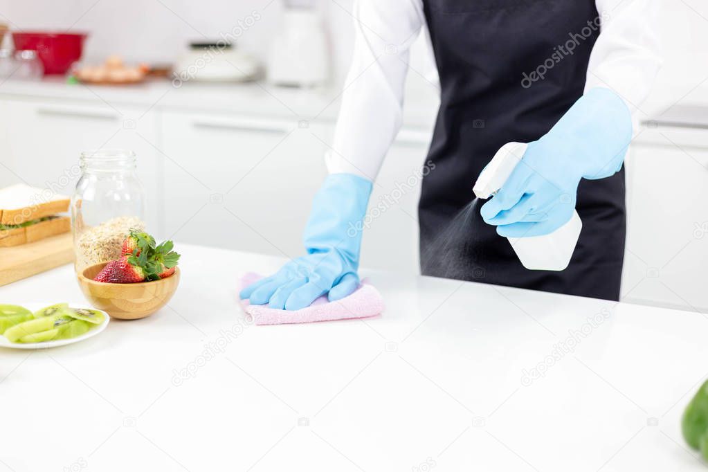 Attractive beautiful woman or housewife is wiping, cleaning and spaying antiseptic or disinfectant on table at kitchen. Cleaning can kill germ, bacteria. Charming beautiful girl wear gloves, apron.