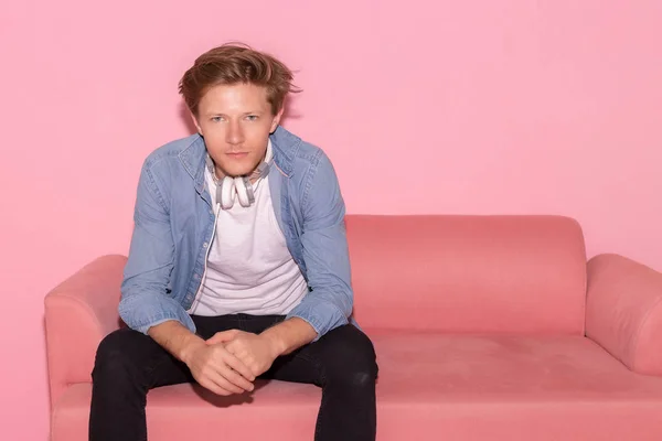 Portrait cool guy. Charming handsome young man looks confident and determined. Attractive handsome guy wear jeans, denim shirt, t shirt. He sits on pink sofa. pink wall background, copy space