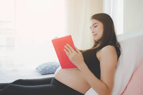 美丽的亚洲妈妈或孕妇读的书，这本书给一些关于产前护理她的婴儿和母亲的知识。母亲坐在卧室的床上。怀孕的女孩得到幸福，放松 — 图库照片