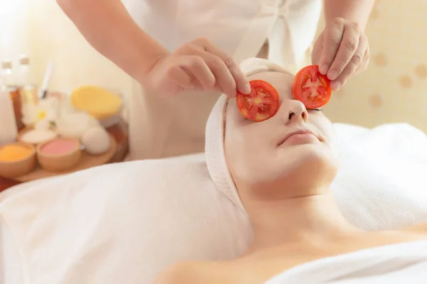 Uma bela jovem deita-se na cama de spa na sala de spa do salão de spa. A massagista pôs partes de tomate em olhos de cliente para fazer a pele de olhos brilhante e refresca. Mulher bonita encantadora obter relaxar, felicidade — Fotografia de Stock