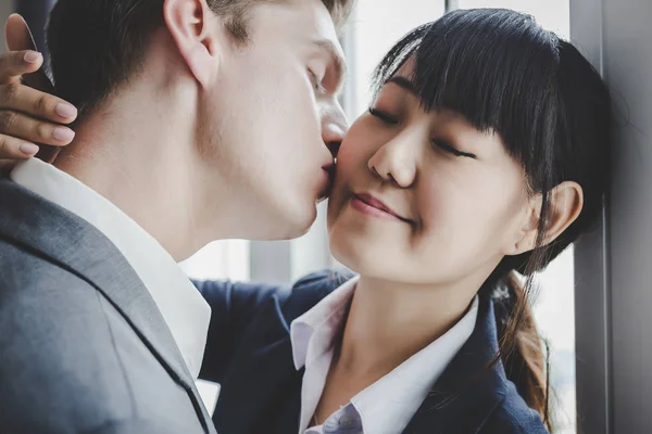 Beautiful young business woman hugging, embracing husband, boyfriend. Handsome young businessman kissing at wife or girlfriend at cheek, affection. Business people falling in love at office. happiness