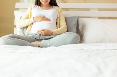 Mutlu hamile anne evdeki rahat yatakta oturarak karnına dokunuyor. Doğum öncesi, hamilelik, annelik, konsept bekleme. Genç hamile kız karnına masaj yapıyor ve fetüsü kontrol ediyor. Boşluğu kopyala