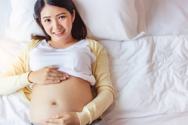快乐怀孕的年轻亚洲女人摸着肚子 带着快乐的微笑在床上赞美她的婴儿或胎儿 年轻的亚洲母亲非常爱她的孩子 妈妈等着要孩子 她的身体很健康 — 图库照片