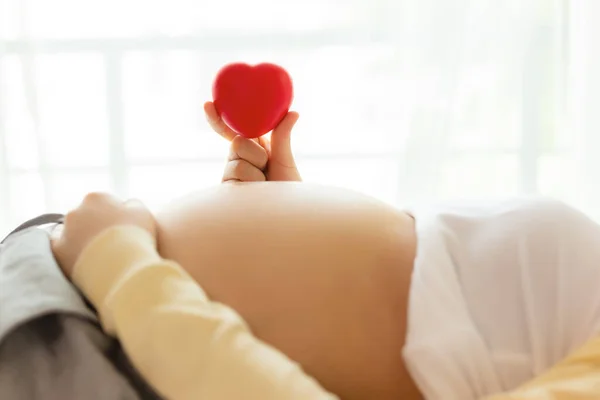 Happy pregnant young woman holding red heart in hand on top of mother belly in hospital bed. prenatal, pregnancy, motherhood, expecting concept. She love baby so much, expecting, waiting give birth