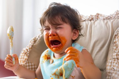 Bebek sandalyesinde yemek yemeyi bitirdiğinde tatminsiz bir şekilde ağlayan sevimli küçük kız ya da bebek bebek. Tatlı kız acıkıyor ve daha fazla yemek istiyor. Karışık ırk kızı kirlenir Çocuk öfkelenir