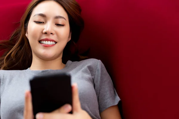在红色沙发上的房子或客厅里用手机描绘快乐的年轻亚洲女人 笑容满面 迷人的女孩与朋友用智能手机发短信 她喜欢5G无线应用 — 图库照片