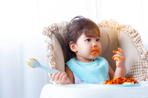 Adorable hungry toddler girl or infant baby eating delicious spaghetti food with tomato sauce on baby chair. Funny cute infant girl get hungry and eating food by herself. Daughter get dirty, look away