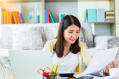 Genç Asyalı kadın evde dizüstü bilgisayarla çalışıyor ve evraklara bakıyor. Oturma odasında gülen yüzle otururken dizüstü bilgisayarda çalışan güzel bir iş kadını. Ev kavramından çalış.