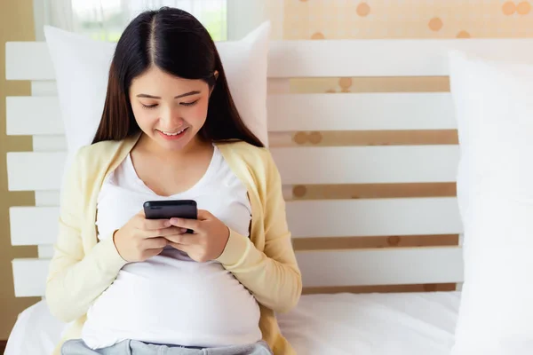 快乐的年轻亚洲孕妇在智能手机上使用和浏览有关产前护理的在线信息 怀孕的母亲坐在家里的床上 技术概念 — 图库照片