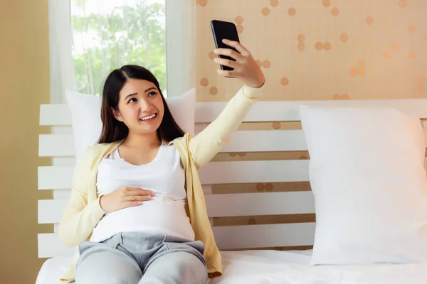 快乐的亚洲孕妇在床上用智能手机自拍 或者年轻的亚洲孕妇在社交媒体上向家人和丈夫展示大肚子 脸上挂着笑脸的视频聊天 — 图库照片