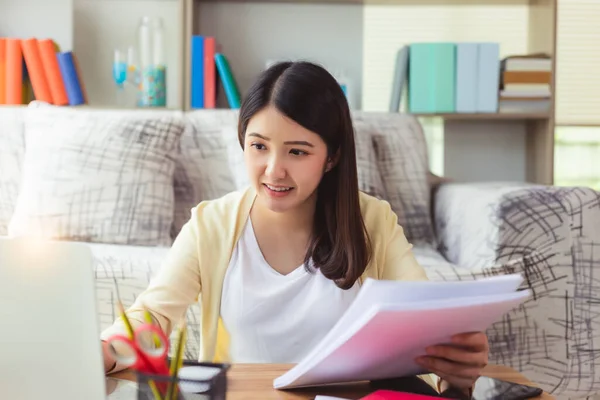 Young asian woman working on notebook computer and holding document at home. Beautiful businesswoman working on laptop computer while sitting at living room with smiley face. Work from home concept.