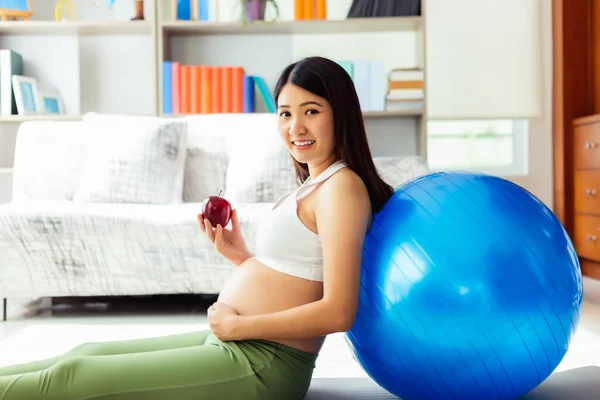 Beautiful young pregnant asian woman holding apple and lean against exercise ball in living room at house with smiley face and looking at camera. Prenatal, pregnancy, motherhood, expectant concept
