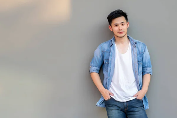 Handsome young Asian man wear denim shirt, looking at camera. Portrait young Asia cool guy with hands in pockets leaning against grey wall. Happy men smiling, confident. He wear jeans. fashion jeans