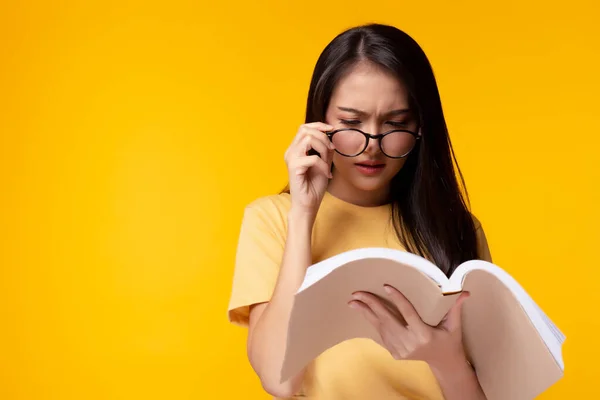 Young woman can not read book because her eyesight\'s getting worse. Young Asian girl wears eyeglasses and holding text book. She is university student. Isolated on yellow background, copy space
