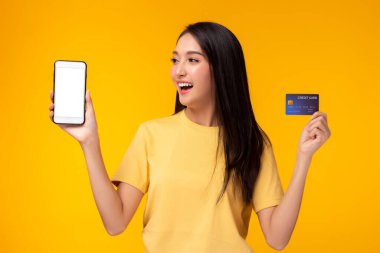 Akıllı telefon ve kredi kartı tutan genç bir kadın ve gülümseyen yüzlü beyaz ekranlı cep telefonuna bakın. Reklamların için kullan. Sarı arka plan. İnternet bankacılığı ve çevrimiçi alışveriş kavramı