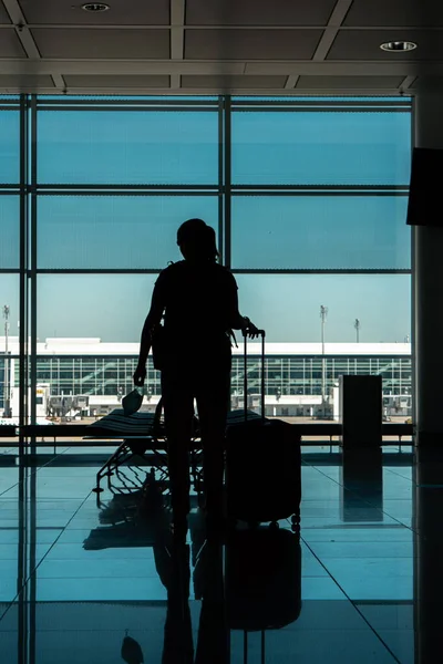 Caucasian young woman silhouette standing in an empty international airport terminal gate with a face mask in her hand next to her roller luggage 2020