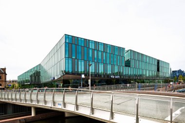 Delft, Hollanda, 26 Ağustos 2025. Ana tren istasyonu modern cam cephe binası. Bulutlu yaz günü, geniş açı, köprüden geçen insanlar.