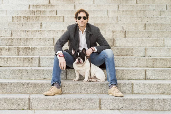 Urban scene. Man dressed in casual clothes sitting on some stairs posing next to his dog