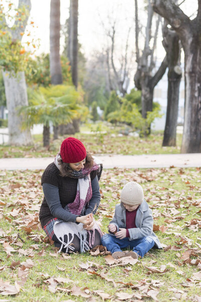 A woman and a child dressed in warm clothes playing with fallen autumn leaves in a park.