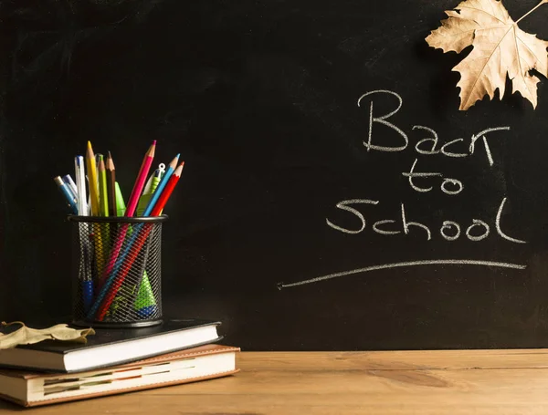 Books, notebooks, school supplies and autumn leaves on wooden boards with a blackboard in the background.