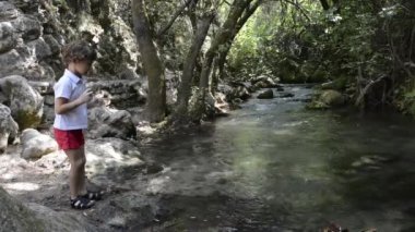 Küçük bir dağ nehrinin kıyısında duran çocuk suya taş atıyor..